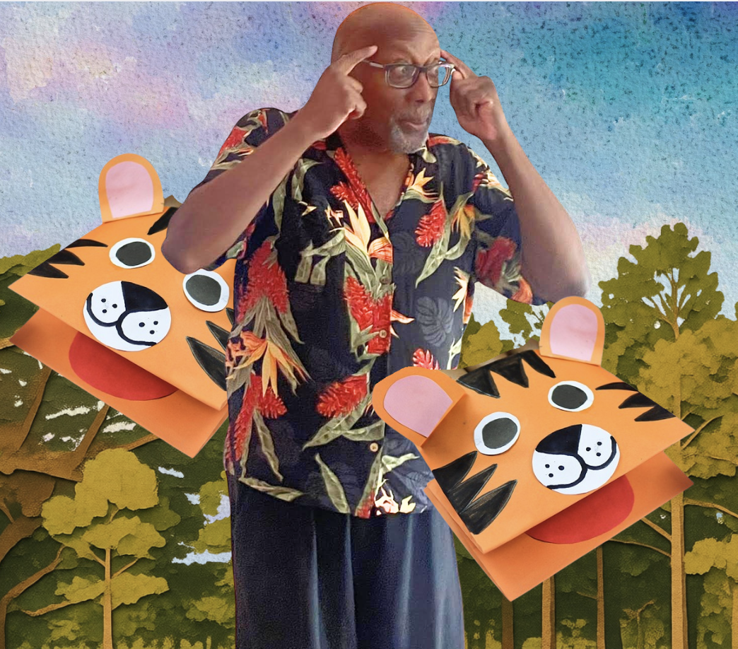 Baden Prince Jr standing beside a painted forest backdrop. He is telling a story. There are two hand made tiger puppets beside him.